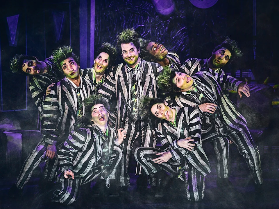 A group of performers in black and white striped suits with green-tinted hair pose together on stage in theatrical lighting and smoky atmosphere.
