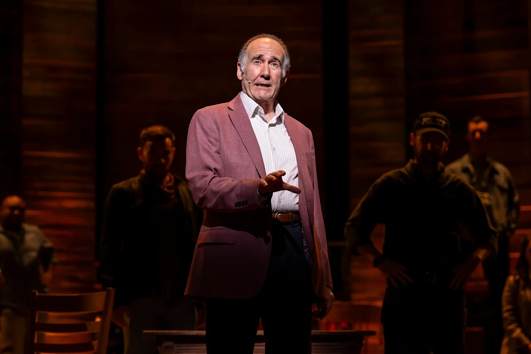 An older man in a pink blazer stands center stage, speaking, with several people in shadow behind him and warm lighting overhead.