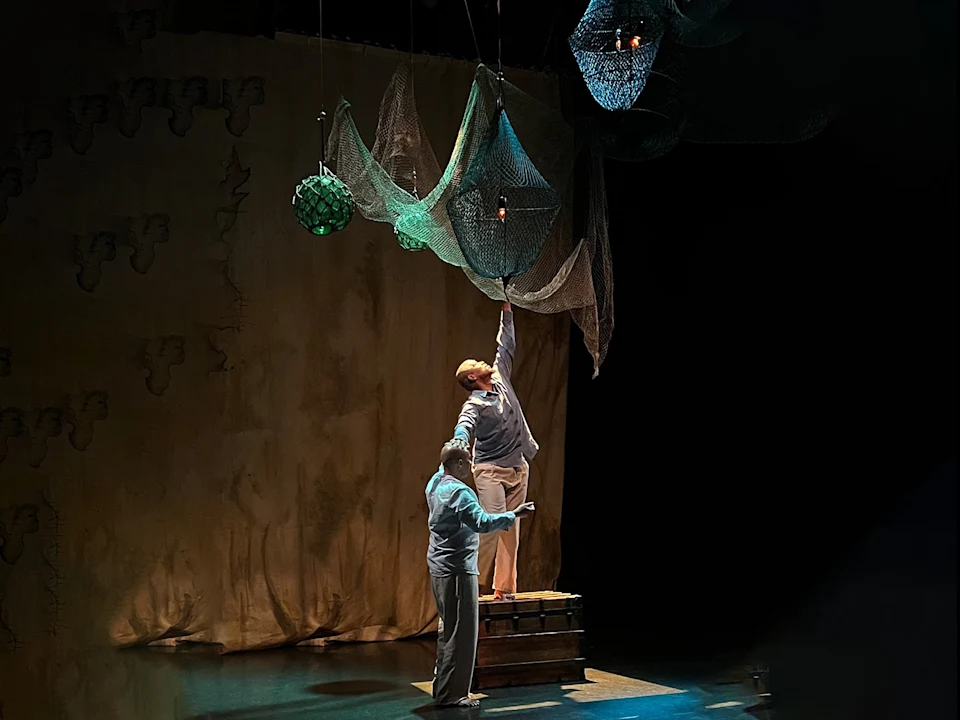 Two actors on stage; one stands on a crate and reaches up toward fishing nets hanging from the ceiling, while the other supports them. The background is dimly lit with a plain curtain.