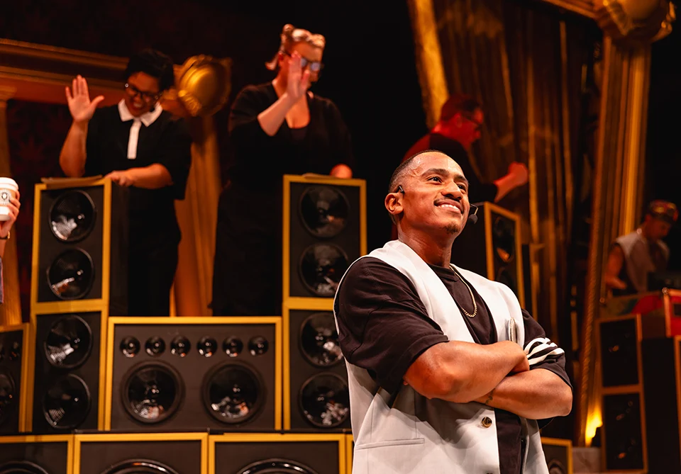 A man stands confidently with arms crossed in front of a wall of speakers, while people in the background wave and interact with the scene.