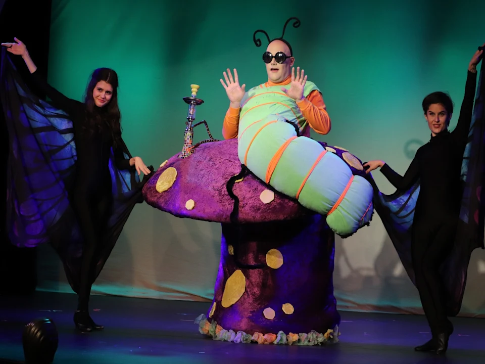 Three actors perform on stage. One in a colorful caterpillar costume sits on a large mushroom prop, while two others in black butterfly costumes pose on either side against a green backdrop.
