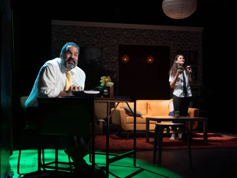 A man sits at a desk with papers and a plant under green light, while a woman stands behind him holding a pen near a couch in a dimly lit room.