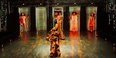 Five women in colorful dresses stand on stage in front of silver doors, with one woman at center front singing or speaking passionately. The floor is reflective and lights line the stage.
