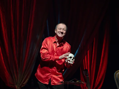 A man in a shiny red shirt stands on stage holding a human skull, smiling, with red curtains in the background and a guitar to the side.