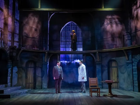 Three actors on stage in a medieval-themed theater set, with one actor on an elevated platform and two actors conversing below. The scene is dimly lit with blue and purple hues.