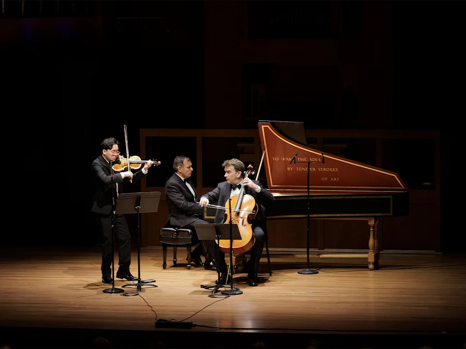 Three musicians perform on stage: one plays violin, one plays cello, and one plays a harpsichord. They are dressed formally and are illuminated under stage lighting.