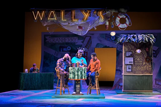 Three men sit at a table under a sign reading "Wally's" on a colorful stage set; a person sits alone at a counter in the background.