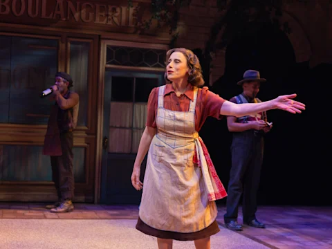 Production photo of The Baker's Wife in New York, showing a woman in an apron gestures with her arm extended onstage, with two men in hats and work clothes standing in the background near a storefront set.