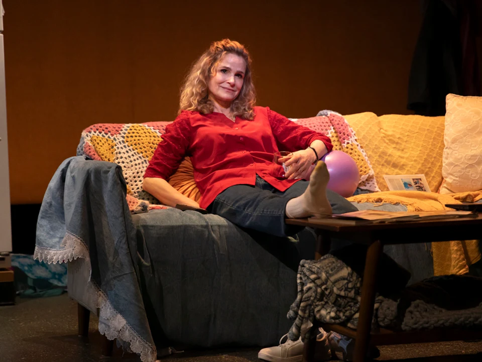 A woman reclines comfortably on a couch with a red blouse and jeans, surrounded by books and a cozy blanket in a warmly lit room.