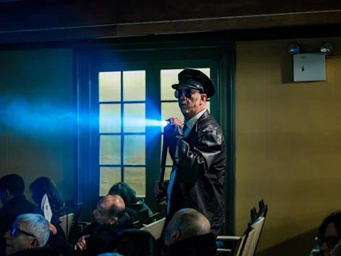 A man in a police uniform and sunglasses shines a bright blue flashlight in a dimly lit room filled with seated people.