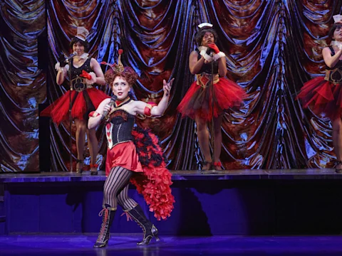 A woman in a red and black burlesque costume sings and gestures at center stage, with three similarly dressed performers singing behind her against a shiny draped backdrop.