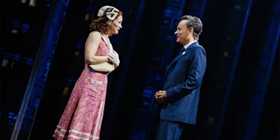 A woman in a pink dress and a man in a blue suit stand facing each other on a stage, engaged in conversation under dramatic lighting.
