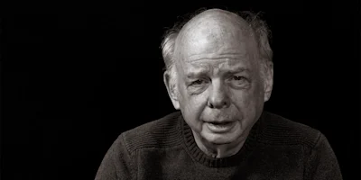 An older man with thinning hair and a serious expression sits against a black background, wearing a dark, textured sweater.