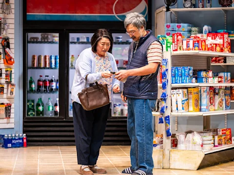 Two adults stand in a grocery store aisle, one handing an item to the other, with shelves of food and drinks visible in the background.