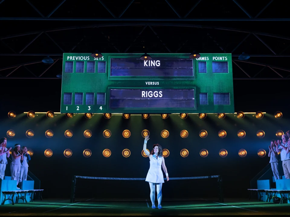 A performer dressed as a tennis player stands center stage under a scoreboard reading "King versus Riggs," with audience members applauding on either side.