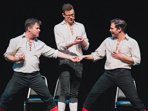 Three men in matching white shirts and black pants perform on stage, with two facing each other in dramatic poses and the third standing behind them, clapping.