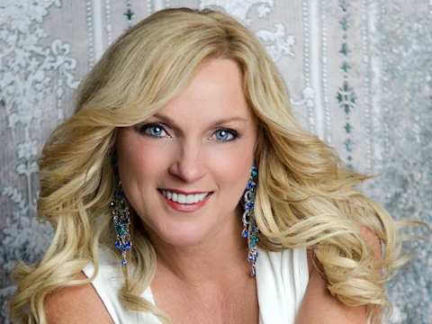 A woman with long blonde hair and blue eyes smiles at the camera, wearing a white top and blue jeweled earrings, with a patterned background behind her.