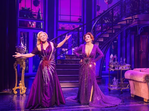 Two women in glamorous purple dresses perform on stage in front of a grand staircase and elegant, purple-lit set.