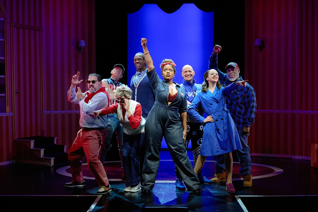 A group of eight actors pose together on a stage with dramatic expressions and gestures, standing in front of a blue backdrop and red-striped walls.