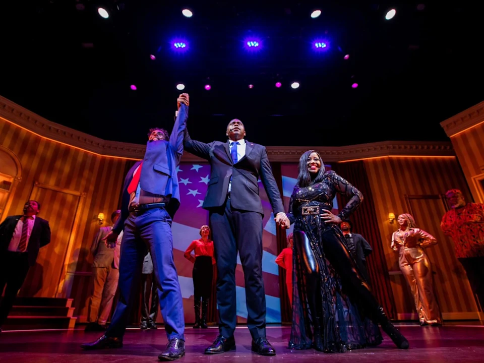A group of performers stands on stage in colorful costumes, with one person in a suit raising another's hand in victory under bright stage lights.