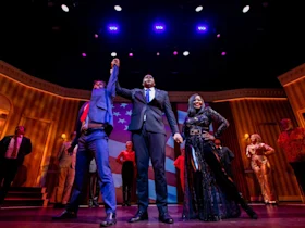 A group of performers stands on stage in colorful costumes, with one person in a suit raising another's hand in victory under bright stage lights.