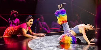 Two performers on stage: one lies on her back with a leg raised, wearing rainbow furry leg warmers; the other kneels nearby, reacting dramatically. Audience members watch in the background.