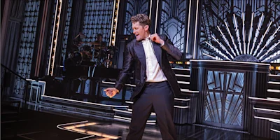 A man in a suit sings and dances on a stage with art deco designs and dramatic lighting, with a band playing in the background.