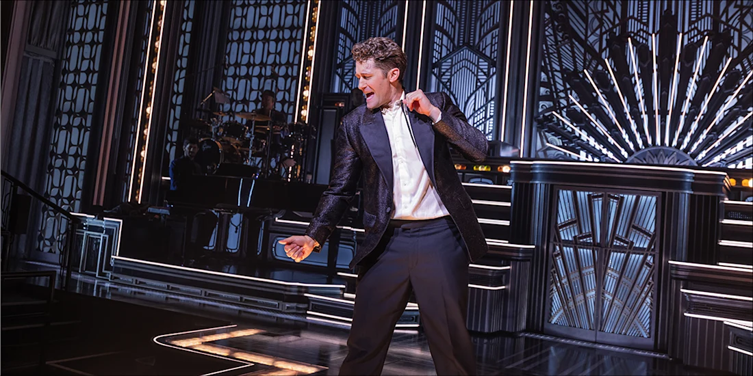 A man in a suit sings and dances on a stage with art deco designs and dramatic lighting, with a band playing in the background.