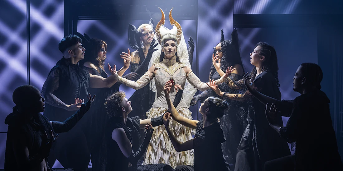 A group of performers in dark costumes surround a central figure in a white, horned costume on a dramatic, smoky stage.