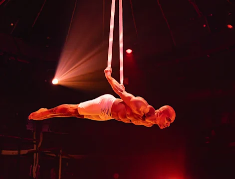 An acrobat performs a suspended pose on aerial straps in a red-lit setting.