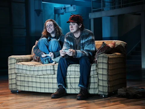 Production photo of Initiative in New York, showing two young people sit on a plaid couch playing video games; one smiles and hugs their knees while the other, wearing a red cap, holds a controller and looks focused.