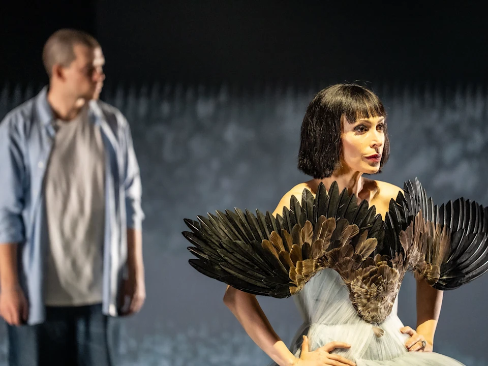 A woman in a dramatic feathered costume stands in focus, while a man in casual clothes is blurred in the background.
