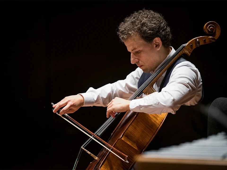 A person wearing a white shirt plays a cello with a bow, focused on the performance, against a dark background.