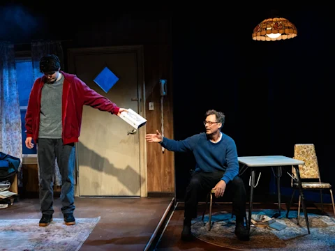 Two men interact in a dimly lit room; one stands holding out a pizza box, while the other sits at a table reaching toward it.