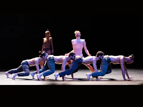 Six dancers on stage, five in a crawling position, one kneeling with arms back, and another standing in the background.