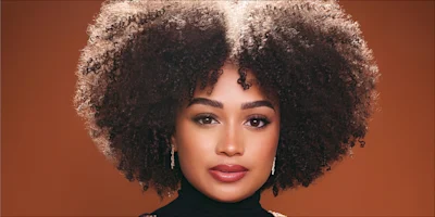 A woman with natural curly hair, wearing a black turtleneck and earrings, looks directly at the camera against a warm brown background.