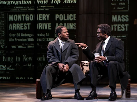 Two men in suits and overcoats sit on a bench, one holding a coffee cup and pointing while talking to the other, with a newspaper headline backdrop behind them.