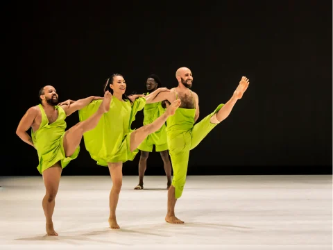 A group of four dancers, dressed in bright green outfits, energetically performing high leg kicks on a stage with a dark background.