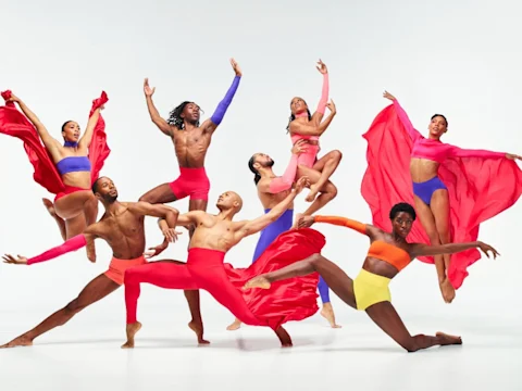 Nine dancers in vibrant red, pink, and orange costumes strike dynamic and expressive poses against a plain white background.