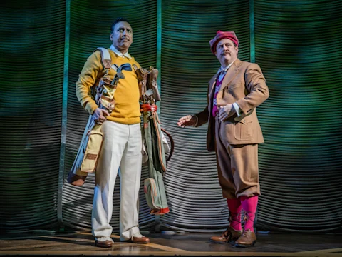 Two men in vintage golf attire stand on stage; one holds multiple golf bags, while the other wears a brown suit with pink socks and a red cap.