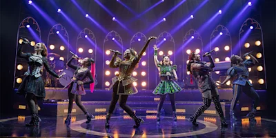 Six female performers in colorful, ornate costumes sing and pose energetically on a stage with bright lights and a dramatic, concert-style backdrop.