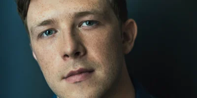 A close-up of a young man with light skin, blue eyes, and short brown hair, looking directly at the camera against a dark background.