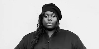 A person wearing a dark beret and buttoned shirt looks directly at the camera against a plain, light-colored background.