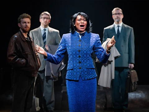 A woman in a blue suit speaks passionately on stage, with three men in suits standing behind her against a dark background.