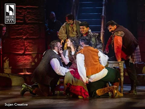 A group of actors in period costumes perform an intense scene around a woman seated at a table on stage, under dramatic lighting.