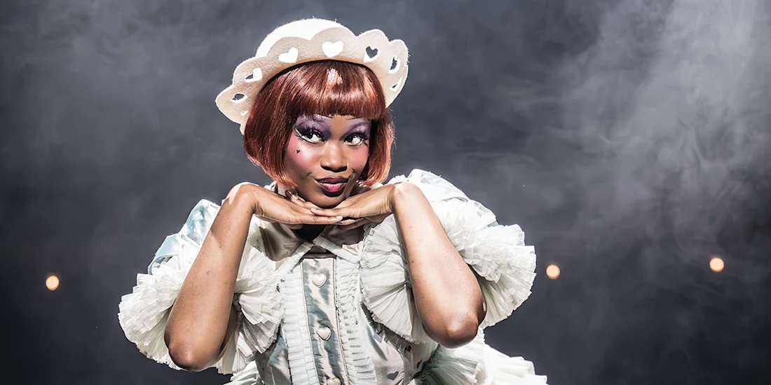 Person in a frilly, light-colored costume and heart-shaped hat poses with hands under chin, against a smoky, dimly lit background.
