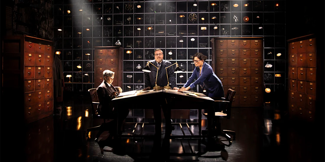 Three people sit and stand around a triangular desk in a dimly lit room with many file cabinets and shelves holding various objects in the background.