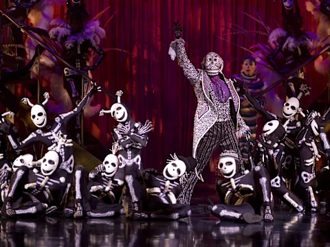 A performer dressed in elaborate skeletal attire stands on a stage with multiple dancers in skeleton costumes posed around them, set against a vibrant red curtain backdrop.