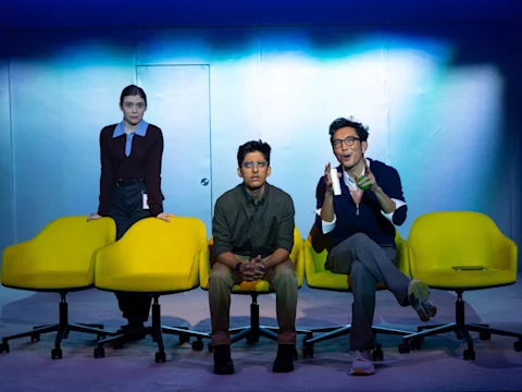 Three people sit or stand beside bright yellow chairs under stage lighting, set against a blue and white backdrop. One gestures while speaking, the others look forward.
