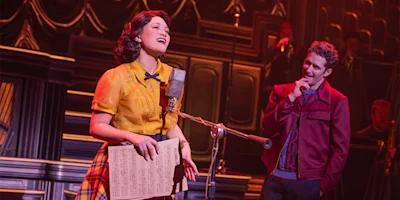 A woman stands at a vintage microphone holding sheet music and singing, while a man in a red jacket watches her on stage.